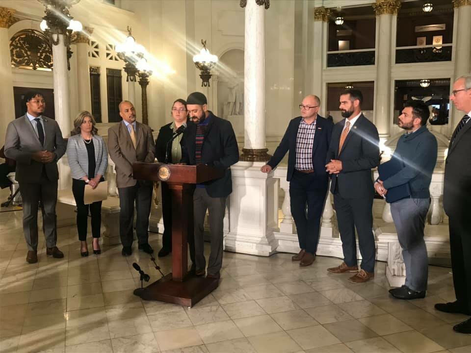 Suited Men at podium announcing the ban bill against Conversion therapy, at Pennsylvania Legislature