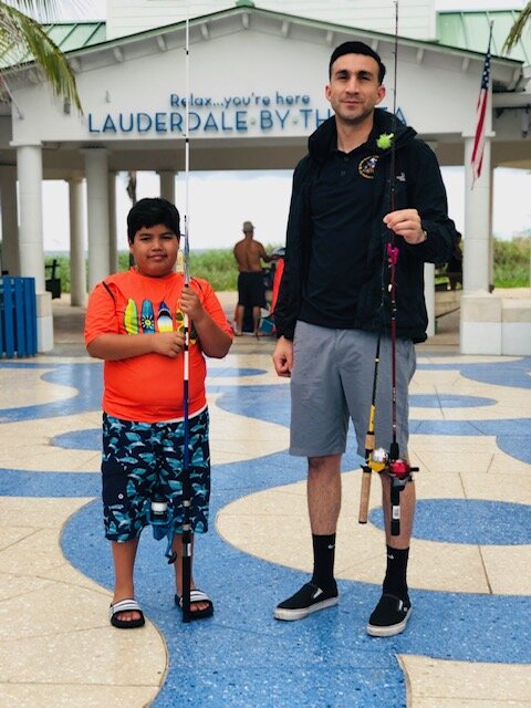 Boy in orange shirt and shorts, and man in black hoodie, both holding fishing poles.