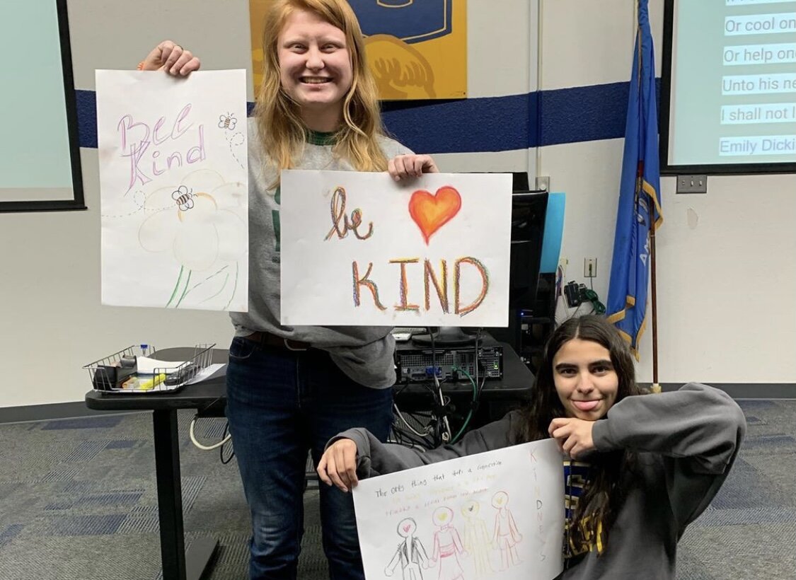 Students in high school celebrating, holding "Be Kind" signs, in classroom.
